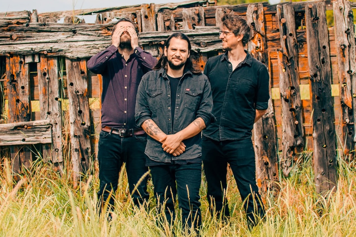 Promo photo of folk band The Longest Johns standing outdoors in tall grass in front of a rustic wooden fence structure.